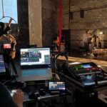 A livestream director and sound operator sit at desks as a theatre event titled Arts for Palestine is livestreamed. theater production lighting, audio and live stream,
