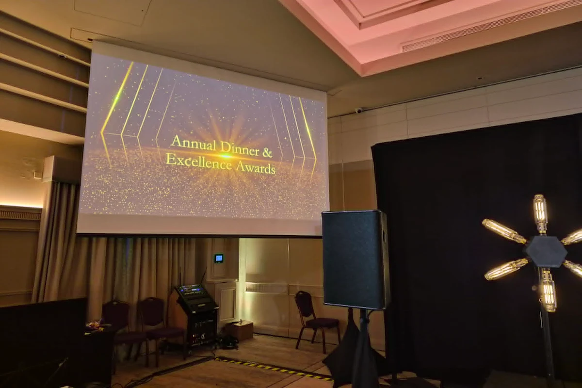 A large projector screen displaying an 'Annual Dinner & Excellence Awards' graphic, supported by professional audio visual services including PA speakers and staging lights inside a corporate event venue